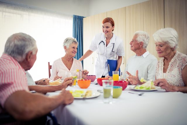 Elderly individuals are enjoying a meal together in a retirement home, accompanied by a nurse who is engaging with them. This image can be used to depict senior living communities, healthcare services, and the importance of social interaction and support for the elderly. Ideal for use in brochures, websites, and advertisements related to elderly care, retirement homes, and healthcare services.