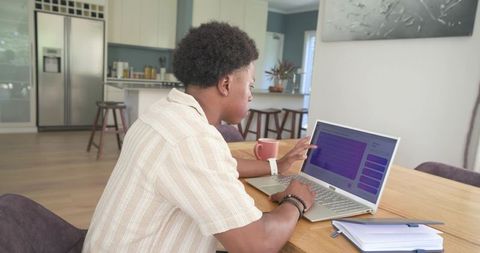 African american man working remotely on laptop at modern kitchen table with coffee mug