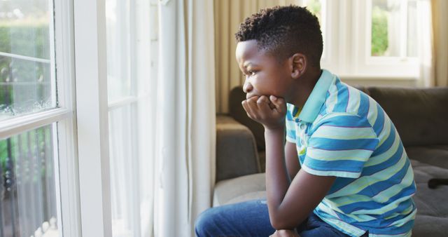 Stressed african american boy looking out of the window while sitting on the couch at home. staying at home in self isolation