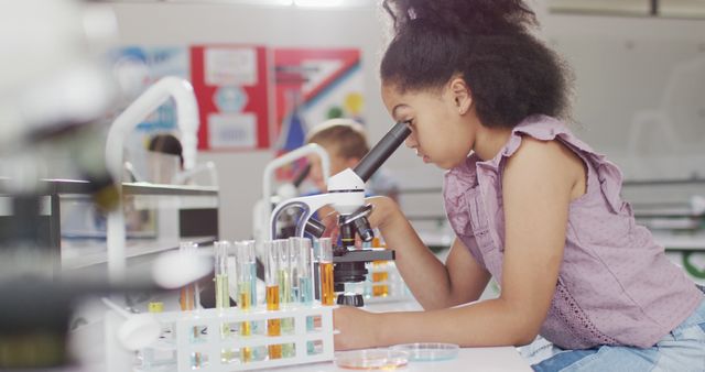 Young girl intently observing samples through microscope during classroom science experiment. Various colorful test tubes are arranged in racks. Ideal for educational materials, STEM programs, classroom activities, and promotional content for science education.