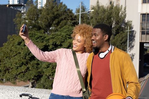 African american couple taking selfie at urban overlook with suitcase and basketball