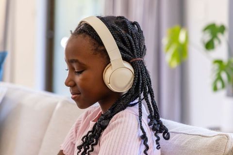 Young african girl listening with cream headphones on sofa, braids, cozy home lifestyle