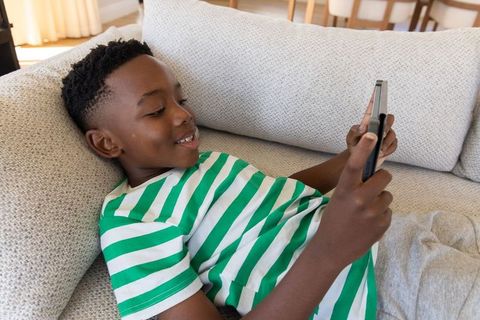 African american boy relaxing on sofa using smartphone for gaming and messaging at home