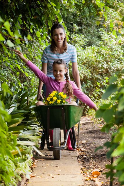 Woman pushing girl sitting with outstretched in wheelbarrow on footpath amidst plants at garden