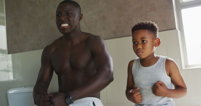 Image of happy african american father and son showing muscles in mirror. Family, spending quality time together at home concept.