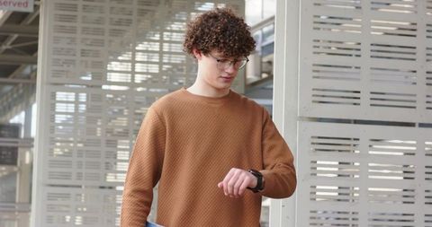 Curly young man checking smartwatch while carrying laptop in urban corridor