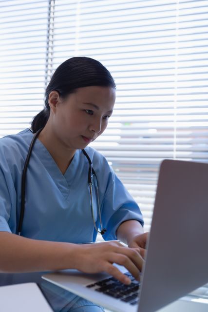 Female surgeon in blue scrubs is working on a laptop while seated at a desk in a clinical setting at a hospital. This image is ideal for use in content related to healthcare, medical professionals, hospital environments, and technological advancements in medicine. Suitable for promotional materials, educational resources, and articles focused on health and medical services.