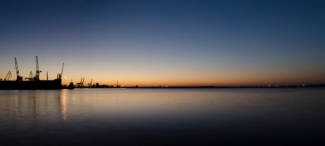 Industrial port at twilight with calm water and silhouettes of cranes against a colorful evening sky. Ideal for use in projects related to maritime industry, logistics, shipping, and tranquil evening landscapes.