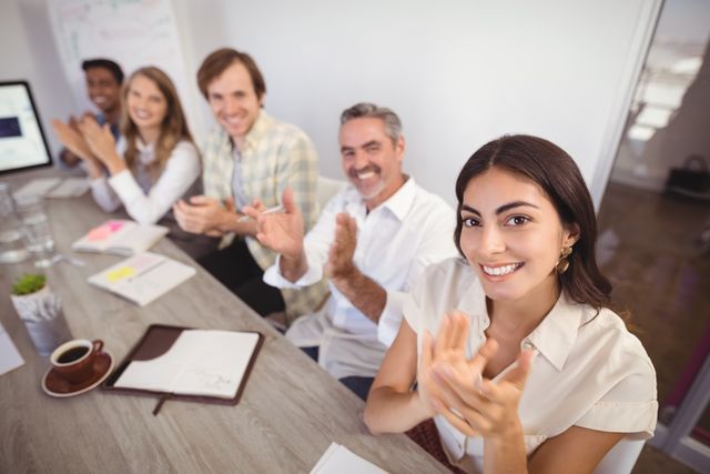 Diverse group of business professionals sitting at a conference table, smiling and applauding. Ideal for use in corporate presentations, teamwork and collaboration themes, business success stories, and professional development materials.