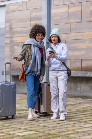 Diverse female friends checking smartphone while traveling with luggage near modern facade