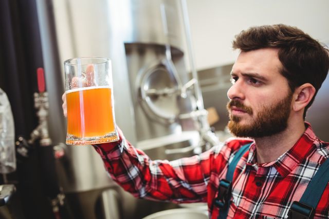Man with beard in plaid shirt examining quality of freshly brewed beer in jug at brewery. Ideal for use in articles about beer production, brewing industry, quality control processes, and craft beer culture. Useful for promoting brewery tours, beer tasting events, or brewing equipment.