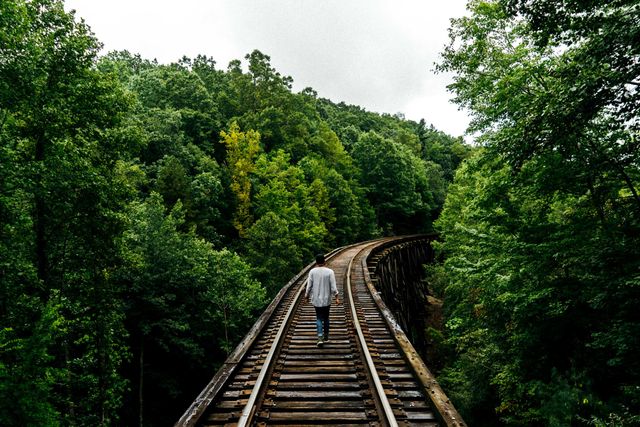 Person walking along wooden railway tracks in dense forest surrounded by extensive greenery. Rugged and nature-filled environment embodies adventure and solitude. Ideal for articles or advertisements focusing on travel, exploration, adventure, hiking, rural retreats, and nature photography.