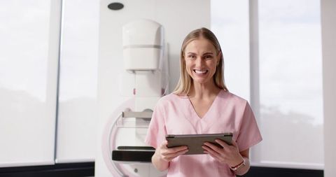 Female radiology technologist smiling while holding tablet beside mammography machine