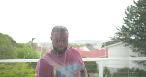 Standing man gazing over suburban rooftops from balcony through streaked glass panel