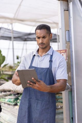 Indian man using tablet in greenhouse nursery wearing denim apron and polo shirt