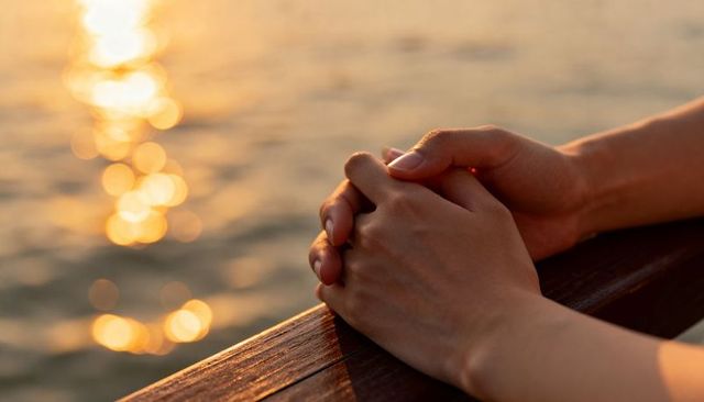 Clasped hands on wooden railing watching golden sunlight reflection on calm water