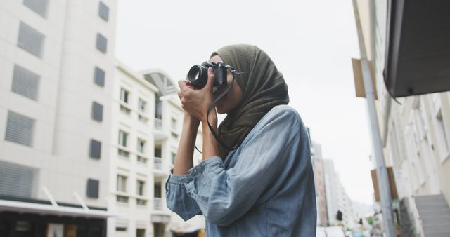Happy biracial woman in hijab taking photos with camera in city street. City living, creativity and modern urban lifestyle.