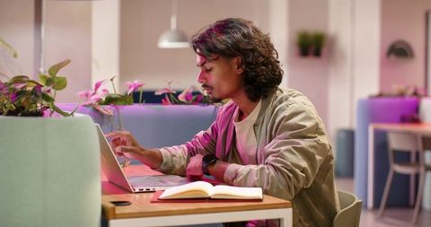 Young man typing on laptop in modern co-working lounge with notebook, plants and pastel lighting
