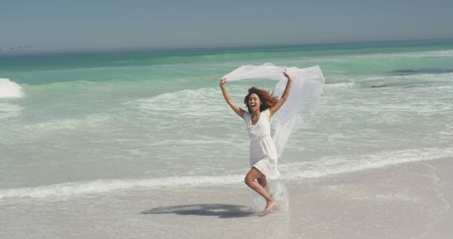 Joyful woman in white dress enjoying the beach with flowing fabric on a sunny day. Captures carefree summer vibes, perfect for travel promotions, vacation themes, and wellness content.