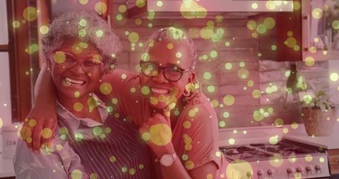 Smiling senior women hugging in cozy kitchen wearing apron and glasses with golden bokeh lights