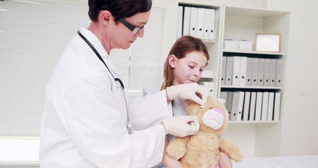 Female doctor demonstrates a medical procedure on a teddy bear to a young female patient in a medical office. This is useful for websites or promotions for pediatric clinics, healthcare providers, education about doctor visits for children, and articles on making children comfortable with medical care. Conveys empathy and patient care in a healthcare setting.
