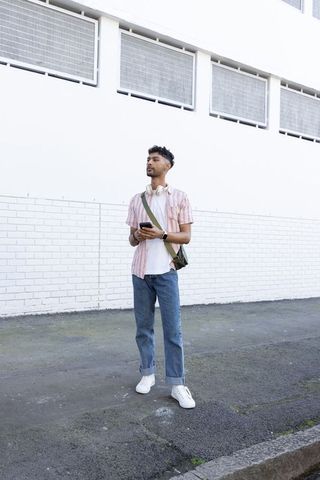 Indian man standing and checking smartphone on urban sidewalk with white brick wall