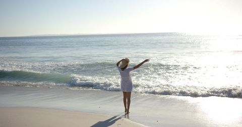 Woman in White Dress Embracing Ocean Breeze on Sunny Beach
