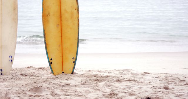 A surfboard standing upright on a sandy beach near gentle ocean waves. Ideal for promoting summer activities, surfing events, coastal travel destinations, or beach-related products.