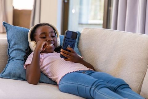 Smiling child relaxing on sofa listening to music with headphones while using smartphone