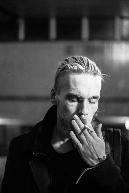 Young man with blond hair smokes a cigarette in an urban setting at night. Black and white photo captures a reflective and contemplative mood. Suitable for use in articles about urban living, nightlife, contemporary youth culture, or smoking habits.