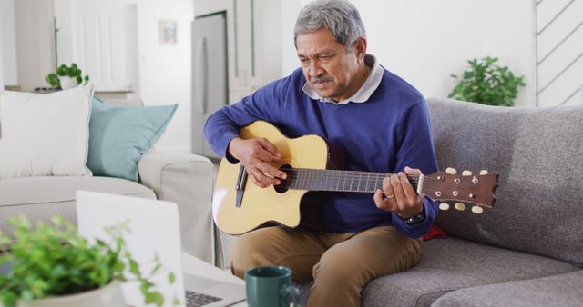 Image of happy senior biracial man using laptop and playing acoustic guitar at home. Retirement, hobbies, global communication, domestic life, inclusivity and senior lifestyle concept.