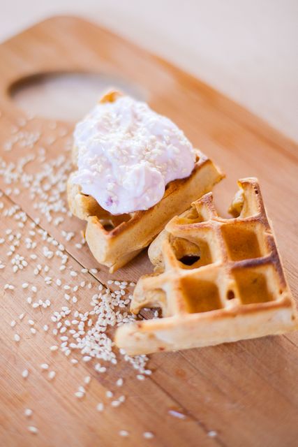 Two waffle pieces topped with cream and sesame seeds are displayed on a wooden board. Ideal for culinary blogs, breakfast menus, recipe magazines, and social media posts focusing on brunch, dessert or homemade bakery products.