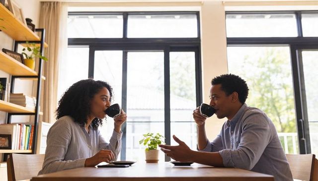 Couple sipping coffee at sunlit dining table, cozy home conversation morning routine lifestyle