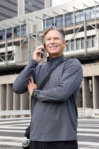 Active senior man talking on phone in modern urban plaza with bag and water bottle