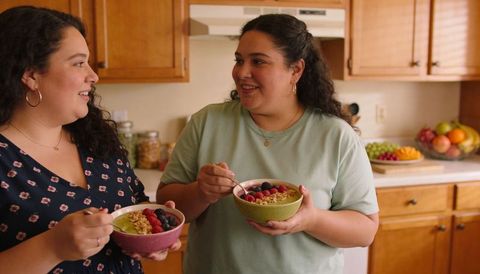 Two women sharing smoothie bowls in cozy kitchen eating granola and fresh berries smiling
