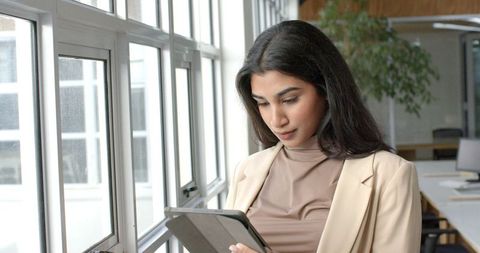 Indian professional using tablet by office windows in beige blazer modern workspace daylight