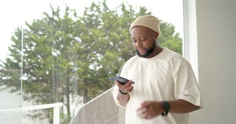 Man checking phone by balcony glass door wearing beanie and casual t-shirt