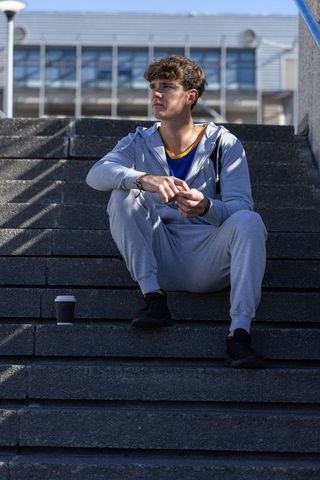 Teen sitting on urban concrete stairs in grey tracksuit holding coffee cup looking pensive