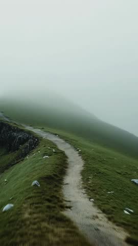 Drone flying along misty ridge trail, advancing on winding dirt path over grassy cliffs