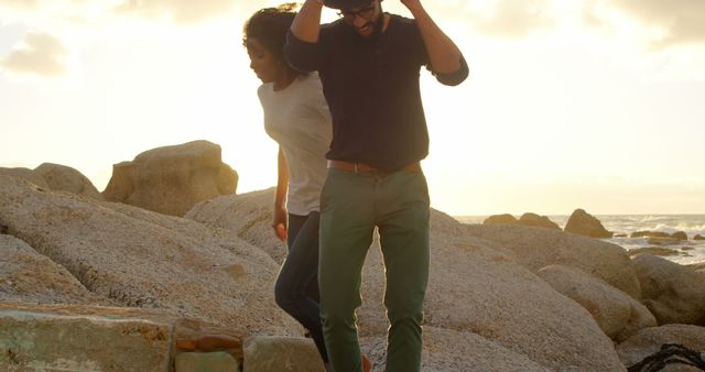 Romantic diverse couple walking on rocks on beach at sunrise, copy space. Summer, vacation, romance, love, relationship, free time and lifestyle, unaltered.