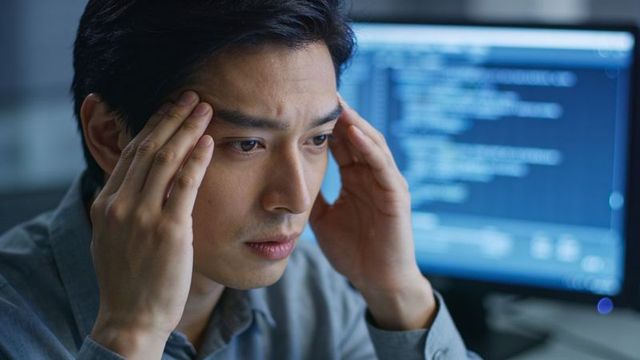 Programmer holding temples and concentrating while debugging code on desktop monitor
