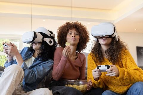 Diverse friends enjoying VR gaming night on sofa sharing snacks, controllers and laughter