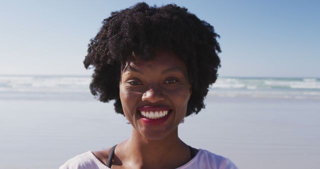 Smiling woman with afro hairstyle enjoying sunny day at the beach. Perfect for use in travel magazines, summer vacation promotions, health and wellness advertisements, or any project focusing on happiness and positivity.
