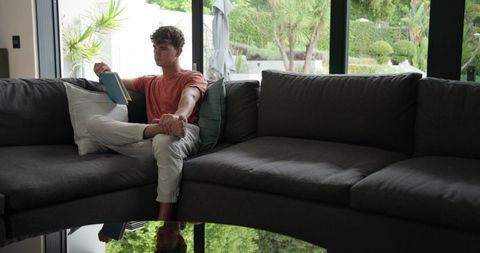 Young man reading paperback on dark sectional sofa by glass doors overlooking lush garden
