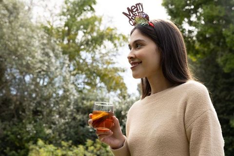 Asian woman wearing merry christmas headband holding citrus drink and smiling in backyard