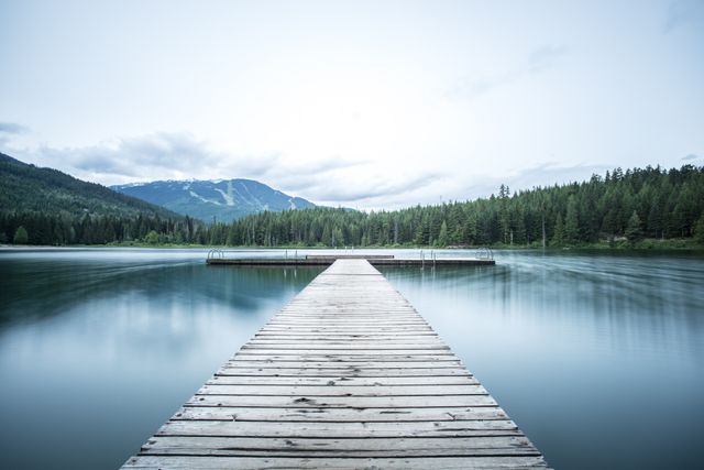 Serene wooden pier extending over a calm mountain lake surrounded by forest and mountains. Tranquil reflections on the water during early morning light. Suitable for use in travel brochures, relaxation themes, wilderness retreats, inspirational posters, nature magazines, and outdoor activity promotions.