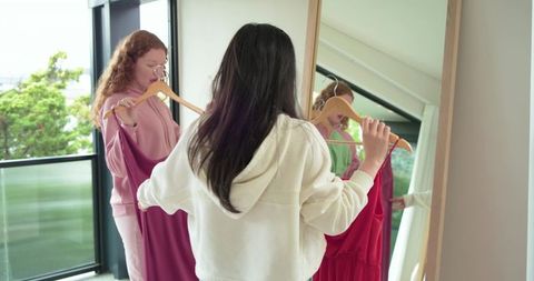 Diverse friends trying on red dress in front of mirror by balcony, casual styling moment