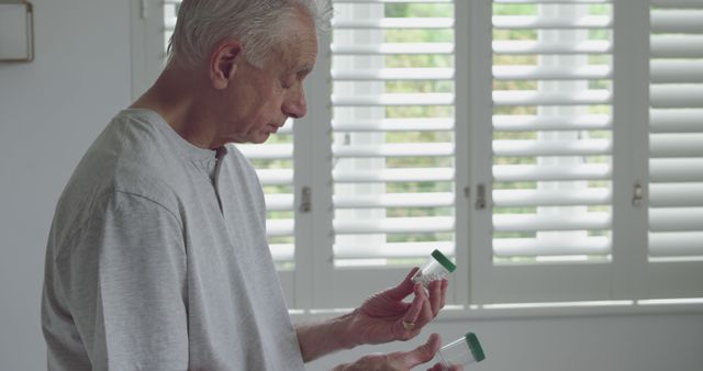 Senior man holding two prescription bottles inside home in front of white shutters. Suitable for use in healthcare, medication management, elderly care, health decision-making, and daily routine content.