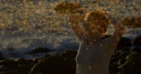 Golden backlit silhouette woman on rocky shore with sea spray bokeh at sunset