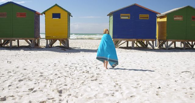 Person wrapped in blue towel walking on white sandy beach near vibrant colorful cabins. Can be used for promoting travel destinations, summer vacations, and coastal activities.